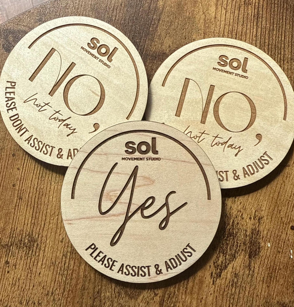Three round wooden movement discs on a wooden surface, featuring laser-engraved text. The front disc is light wood with dark brown engraving that reads 'sol MOVEMENT STUDIO', 'Yes', and 'PLEASE ASSIST & ADJUST'.